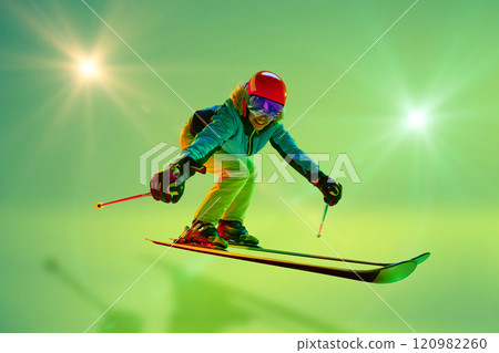 Low angle, side view of female athlete, skier in colorful gear mid-air jump neon green background. Dynamic pose. 120982260