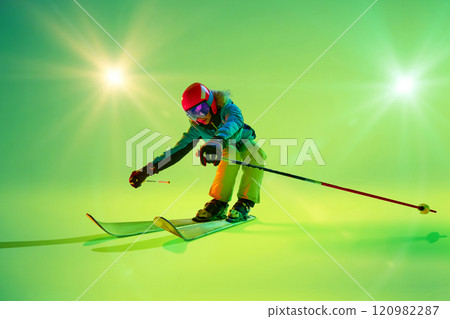 Captured in mid-motion, female skier skiing as on downhill and skier's reflective goggles shimmer under bright neon lights, against studio background. Captured in mid-motion, female skier skiing as on downhill and skier's reflective goggles shimmer under bright neon lights, against studio background. 120982287