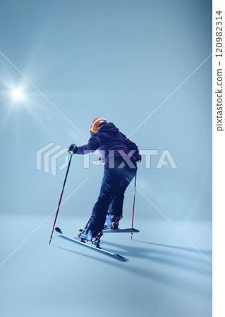 Athletic woman skier in purple jacket leans into precise carve, their skis cutting smoothly against gradient blue studio background. Athletic woman skier in purple jacket leans into precise carve, their skis cutting smoothly against gradient blue studio background. 120982314
