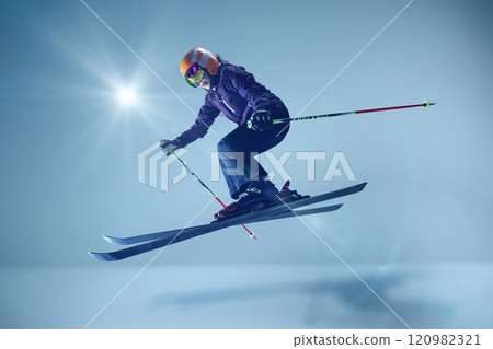Winter athlete launching dynamically, orange helmet and reflective goggles glinting under studio lighting, emphasizing speed against seamless blue. 120982321