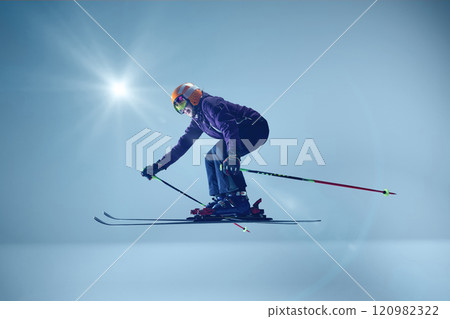Side view of athlete executing precise ski jump, wearing purple jacket and mirrored goggles against blue backdrop and sunlight enhancing action Side view of athlete executing precise ski jump, wearing purple jacket and mirrored goggles against blue backdrop and sunlight enhancing action 120982322