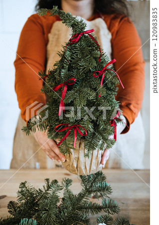 Woman holding handcrafted mini Christmas tree decorated with red ribbons. Promotes eco-friendly holiday decor and sustainable Christmas trends. Woman holding handcrafted mini Christmas tree decorated with red ribbons. Promotes eco-friendly holiday decor and sustainable Christmas trends. 120983835
