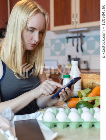A woman checks the calorie content of food in a smartphone app. Calorie counting for weight loss. There are products for dieting on the table 120984360