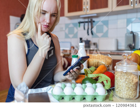 A woman checks the calorie content of food in a smartphone app. Calorie counting for weight loss. There are products for dieting on the table A woman checks the calorie content of food in a smartphone app. Calorie counting for weight loss. There are products for dieting on the table 120984361