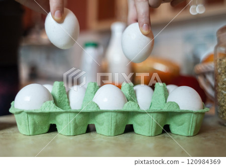 Close-up of eggs in a green box. A woman picks up an egg with her fingers from a container to cook a dish in her kitchen 120984369
