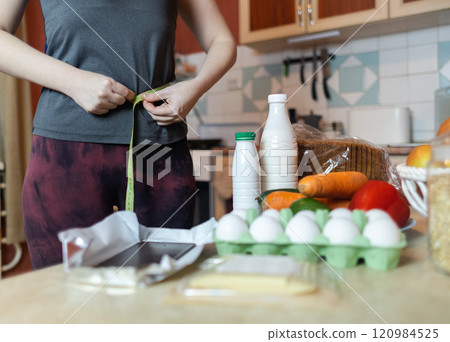 A woman checks her belly circumference with a measuring tape. There are diet foods on the table. Healthy lifestyle and weight loss A woman checks her belly circumference with a measuring tape. There are diet foods on the table. Healthy lifestyle and weight loss 120984525