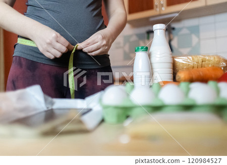 A woman checks her belly circumference with a measuring tape. There are diet foods on the table. Healthy lifestyle and weight loss A woman checks her belly circumference with a measuring tape. There are diet foods on the table. Healthy lifestyle and weight loss 120984527