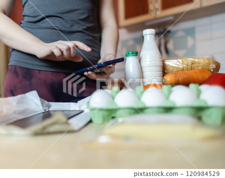 A woman checks the calorie content of food in a smartphone app. Calorie counting for weight loss. There are products for dieting on the table 120984529