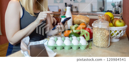A woman checks the calorie content of food in a smartphone app. Calorie counting for weight loss. There are products for dieting on the table A woman checks the calorie content of food in a smartphone app. Calorie counting for weight loss. There are products for dieting on the table 120984541