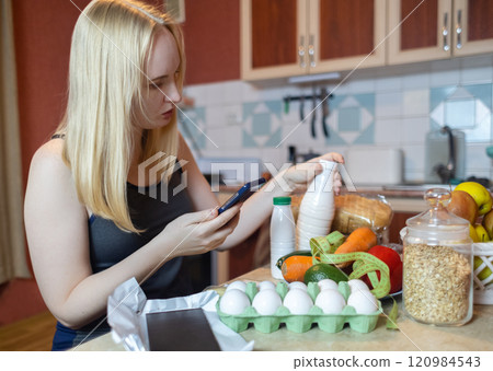 A woman checks the calorie content of food in a smartphone app. Calorie counting for weight loss. There are products for dieting on the table 120984543