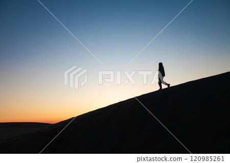 Silhouette of a woman walking on a sand dune at sunset, under a tranquil and colorful evening sky. Capturing happy, peaceful summer vacation on beach. Silhouette of a woman walking on a sand dune at sunset, under a tranquil and colorful evening sky. Capturing happy, peaceful summer vacation on beach. 120985261