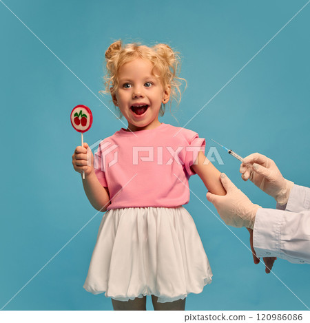 Smiling happy little girl, child in cute outfit, holding lollipop candy while doctor making vaccine injection on her arm against blue studio background Smiling happy little girl, child in cute outfit, holding lollipop candy while doctor making vaccine injection on her arm against blue studio background 120986086