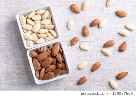 Almonds in square shaped white bowls, on linen fabric. Almonds with seed coats and blanched. Fruits of Prunus amygdalus, syn. Prunus dulcis, ready-to-eat as snack or as ingredient for baking. Photo. 120987699