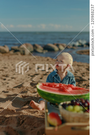 Baby toddler in blue hoodie sits on beach next to picnic with watermelon and fruit on sunny day Baby toddler in blue hoodie sits on beach next to picnic with watermelon and fruit on sunny day 120988237