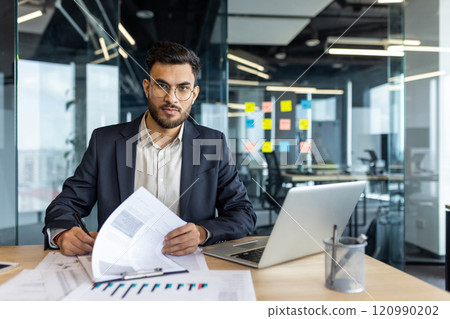 Businessman in a modern office holding documents and working on a laptop. The setting is professional, emphasizing focus and readiness in a business environment. Ideal for corporate themes. Businessman in a modern office holding documents and working on a laptop. The setting is professional, emphasizing focus and readiness in a business environment. Ideal for corporate themes. 120990202