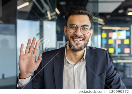 A confident businessman in a modern office environment participates in a video call, looking directly at the camera. He waves, conveying professionalism and friendliness, dressed in a formal suit 120990272