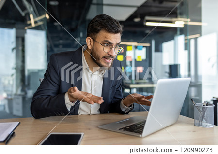 An exasperated businessman in an office uses a laptop for a video call, expressing frustration. His smartphone is visible, adding to the tech-focused environment. 120990273
