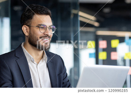 A confident businessman is smiling as he works on his laptop in a bright office setting. His expression conveys professionalism and focus in a modern and productive corporate environment. 120990285