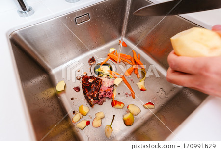 Peeling potatoes over a kitchen sink filled with food scraps and running water. 120991629