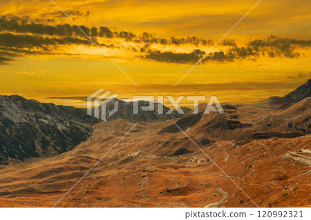 Golden sunset over a majestic mountain landscape with dramatic sky and rocky terrain. 120992321