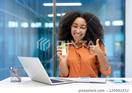 Young woman businesswoman showing pleasure from using a throat spray at her workplace. She is smiling confidently, giving a thumbs up, and sitting at a desk with a laptop and phone Young woman businesswoman showing pleasure from using a throat spray at her workplace. She is smiling confidently, giving a thumbs up, and sitting at a desk with a laptop and phone 120992852