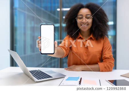 Young woman businesswoman displaying a smartphone at her desk in a modern office environment. She is wearing glasses and smiling, creating a professional yet approachable atmosphere. 120992868