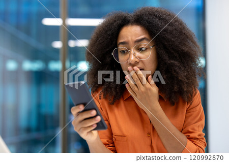 A young businesswoman in an office setting reacts with surprise while looking at her phone. Her expression conveys a sense of shock or unexpected news. A young businesswoman in an office setting reacts with surprise while looking at her phone. Her expression conveys a sense of shock or unexpected news. 120992870