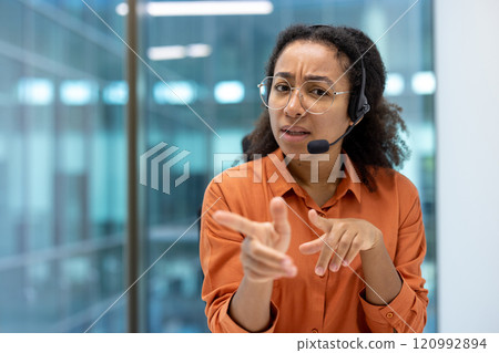 A woman wearing glasses and headset gestures during video call in an office. She appears confident and engaged, looking directly at the camera communicating effectively in a professional environment. 120992894