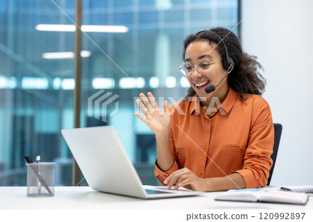A young businesswoman engages in a video call at her desk, using a laptop and wearing a headset. She appears friendly and professional, embodying remote work and communication. A young businesswoman engages in a video call at her desk, using a laptop and wearing a headset. She appears friendly and professional, embodying remote work and communication. 120992897