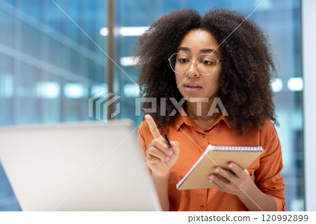 Young woman businesswoman engaged in online learning, attentively watching a video course. She holds a notebook and pen, intensely focused on her laptop screen, absorbing information. Young woman businesswoman engaged in online learning, attentively watching a video course. She holds a notebook and pen, intensely focused on her laptop screen, absorbing information. 120992899