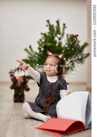 Cute little child girl reads a book near the Christmas tree at home 120992992