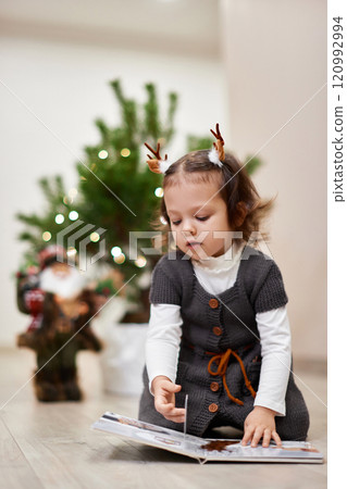 Cute little child girl reads a book near the Christmas tree at home Cute little child girl reads a book near the Christmas tree at home 120992994