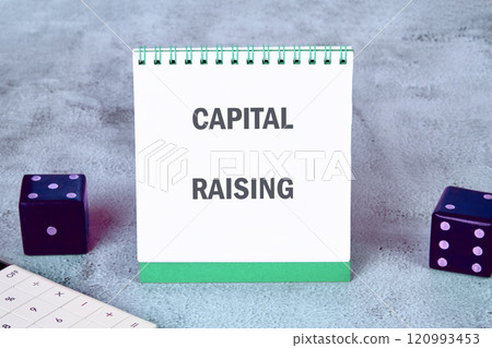 Notepad displaying capital raising concept alongside dice on a textured surface 120993453