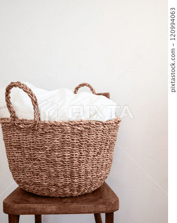 A natural woven basket with white linens inside, placed on a rustic wooden chair against a neutral white wall. A natural woven basket with white linens inside, placed on a rustic wooden chair against a neutral white wall. 120994063