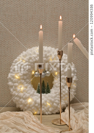 Christmas decor, white soft plush wreath with lights, lit candles in golden holder, beige cloth on wooden table Christmas decor, white soft plush wreath with lights, lit candles in golden holder, beige cloth on wooden table 120994386