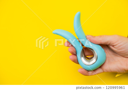 Female hand holding blue vibrator sticking out of yellow paper background.  120995961
