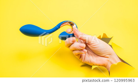 Female hand holding blue vibrator sticking out of yellow paper background. Copy space. 120995963