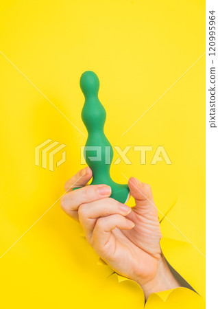 Female hand with green anal plug sticking out of yellow paper background. 120995964