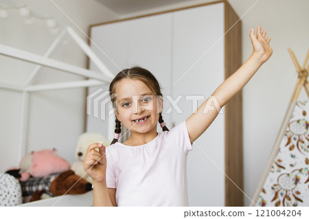 Little Caucasian girl showing missing baby tooth with happy expression in bedroom. Child excited and proud about milestone, holding tooth with raised arm in playful room. 121004204