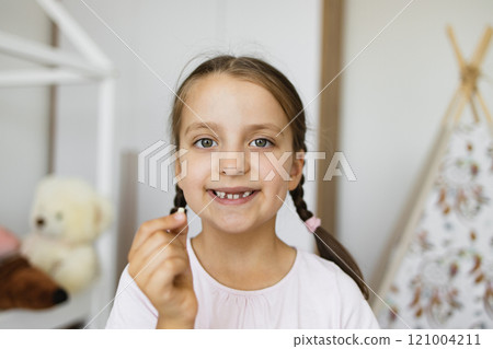 Joyful little Caucasian girl holding lost baby tooth at home, showing excitement and pride. Captured in cozy bedroom setting highlighting this childhood milestone with cheerful smile and braided hair. 121004211