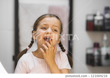 Caucasian girl with braided hair examines her loose tooth and missing teeth in bathroom mirror. Young child with tooth gap experiences curiosity and wonder during dental milestone. Caucasian girl with braided hair examines her loose tooth and missing teeth in bathroom mirror. Young child with tooth gap experiences curiosity and wonder during dental milestone. 121004216