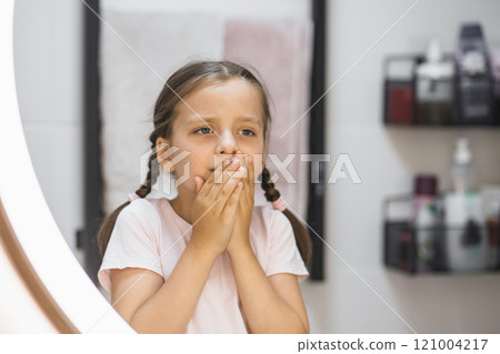 Little Caucasian girl with toothache holds mouth while looking in mirror. Expression of discomfort in bathroom setting. Little Caucasian girl with toothache holds mouth while looking in mirror. Expression of discomfort in bathroom setting. 121004217