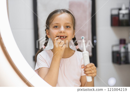 Little Caucasian girl with missing baby teeth using electric toothbrush and smiling in bathroom mirror, showing confidence and dental care. 121004219