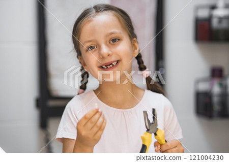 Little Caucasian girl holding a fallen baby tooth with pliers, looking in bathroom mirror. She smiles, showing missing tooth, representing childhood milestones and dental health. Little Caucasian girl holding a fallen baby tooth with pliers, looking in bathroom mirror. She smiles, showing missing tooth, representing childhood milestones and dental health. 121004230