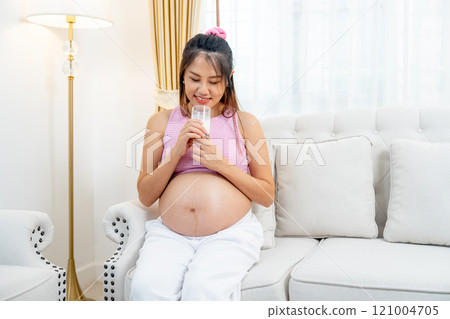 Asian pregnant woman sit on sofa and hold a glass of milk and touch her belly also look at the glass with happiness. 121004705