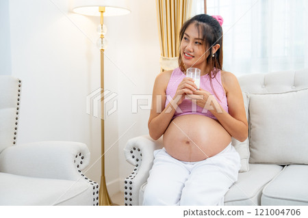 Asian pregnant woman sit on sofa and hold a glass of milk and touch her belly also look to left side with happiness. 121004706