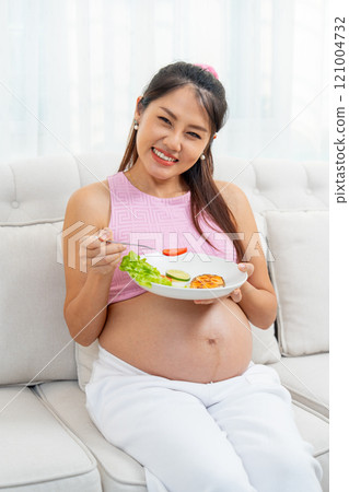 Vertical image of Asian pregnant woman sit on sofa and hold food in disk and use fork pick tomato up and show to camera with smiling. 121004732