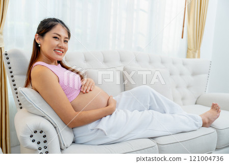 Side view of pregnant woman sit and relax on sofa in living room and look at camera with smiling. Side view of pregnant woman sit and relax on sofa in living room and look at camera with smiling. 121004736