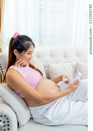 Vertical image of Asian pregnant woman lie on sofa and use tools for listen sound in her belly and look happy at home. Vertical image of Asian pregnant woman lie on sofa and use tools for listen sound in her belly and look happy at home. 121004740