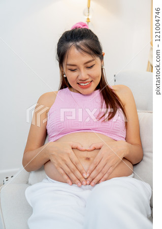 Vertical image of Asian pregnant woman sit on sofa and put hands on her belly with heart shape and smiling. Vertical image of Asian pregnant woman sit on sofa and put hands on her belly with heart shape and smiling. 121004746
